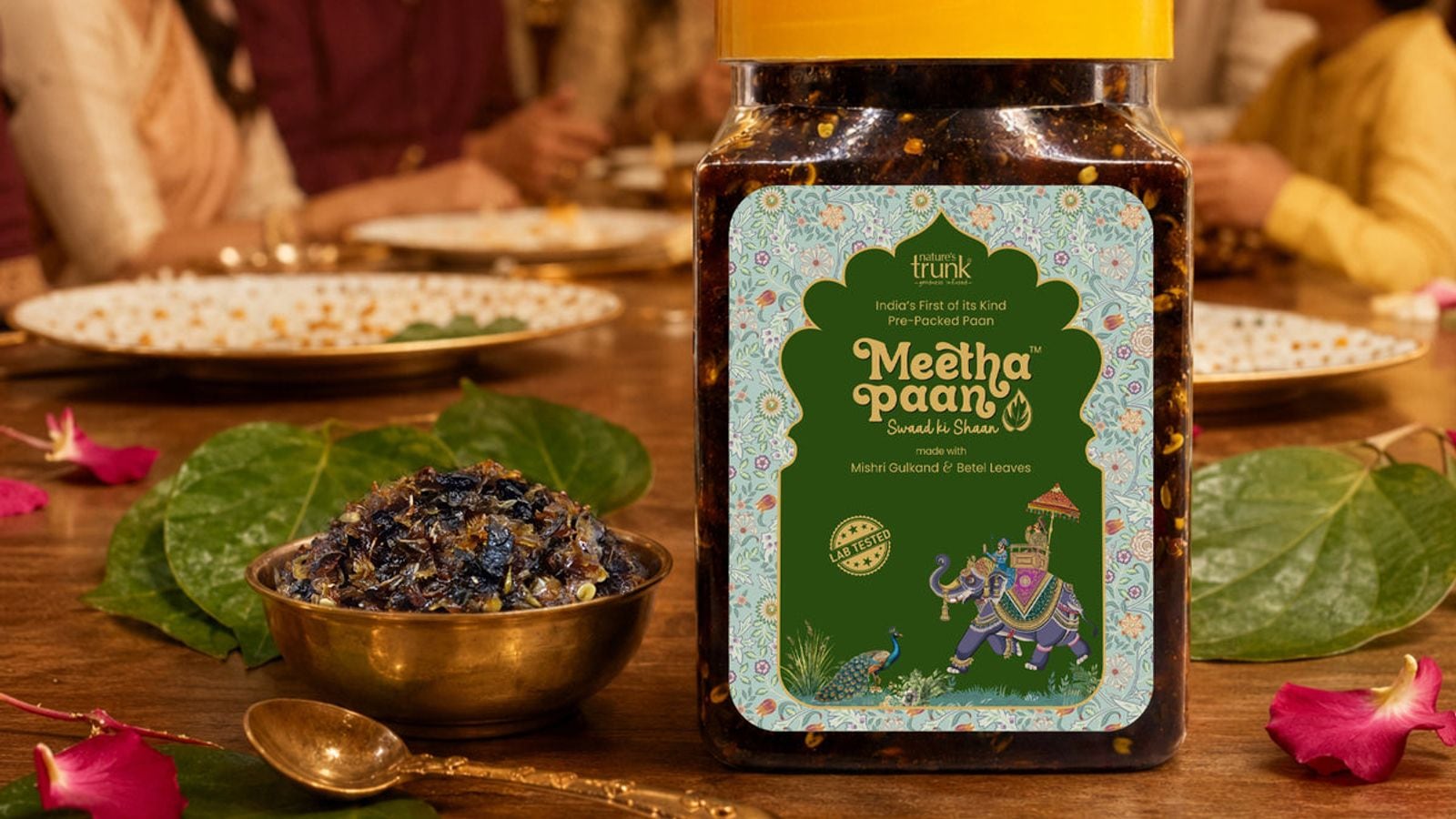 A jar of Nature’s Trunk Meetha Paan on a dining table beside a brass bowl of Meetha Paan, betel leaves, fennel seeds and rose petals